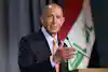 A formally dressed individual speaks at a podium with an Iraqi flag visible in the background.