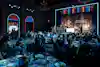 a group of people sitting at tables in a room with a stage and a stage with a stage and a stage with a stage with a stage with a stage with a stage with a stage with a
