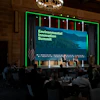 Two individuals engage in a discussion on a stage at the Environmental Innovation Summit, with an audience seated at round tables in a grand hall.