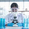 a woman wearing goggles and a white coat looking through a microscope