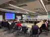 a group of people sitting at a table in a room with a large screen