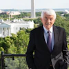 a man in a suit standing on a balcony with trees and a monument in the background
